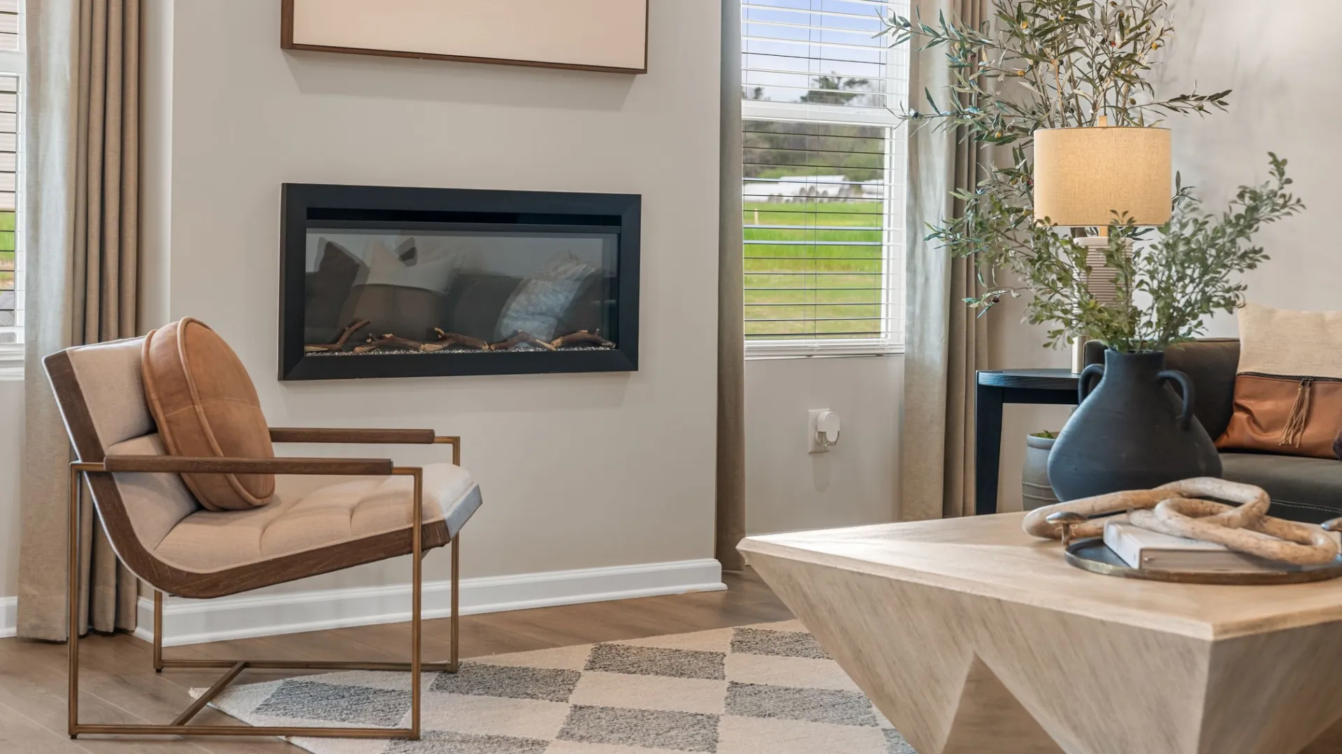 Modern living room with beige armchair, wall-mounted fireplace, wooden coffee table, and large window with blinds.